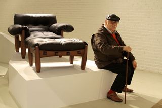 Designer Sergio Rodrigues - seen here with his trademark handlebar moustache, flat cap and Jacaranda wood walking cane - posing next to his Poltrona Mole chair