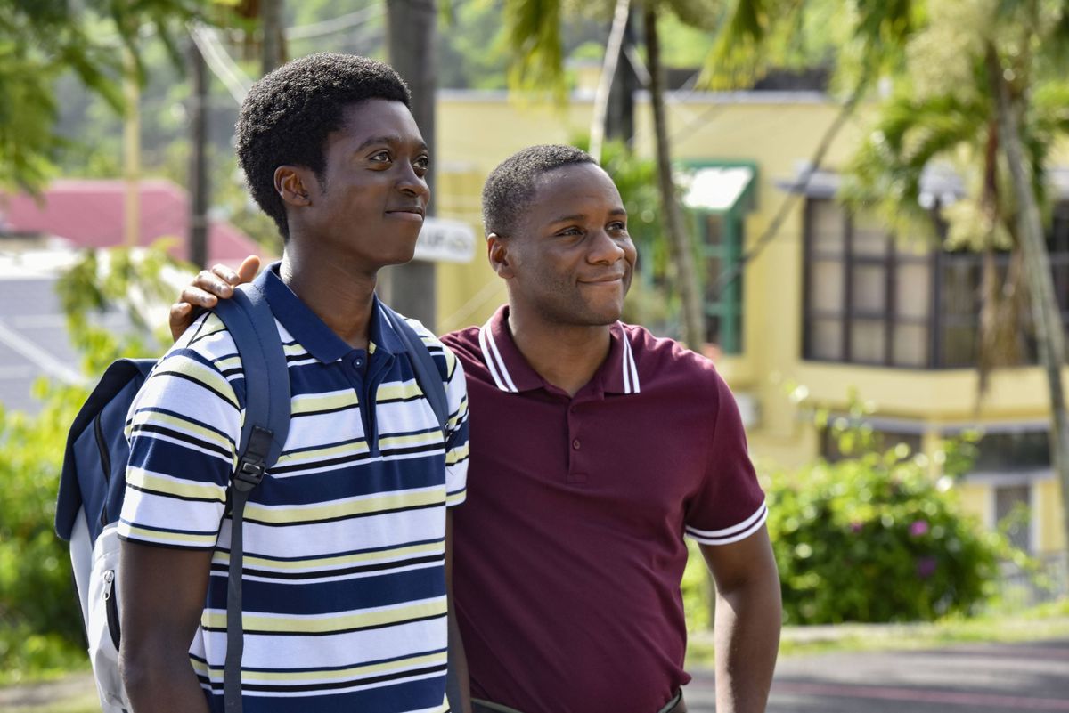 Officer Benjamin Brice (Anthony J Abraham) stands with Sergeant JP Hooper (Tobi Bakare) in Saint Marie, just outside the police station (not visible in the picture). They are both in plain clothes: Benjamin is wearing a blue, white and yellow horizontal striped polo shirt and a blue backpack, and JP is in a red polo shirt. JP has his arm around Benjamin&#039;s shoulder.