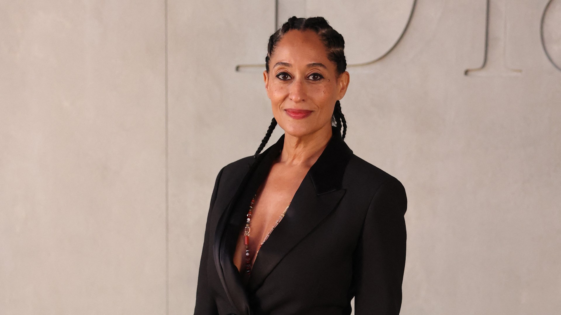 a black woman wears a black blazer and cornrows while posing in front of a white wall