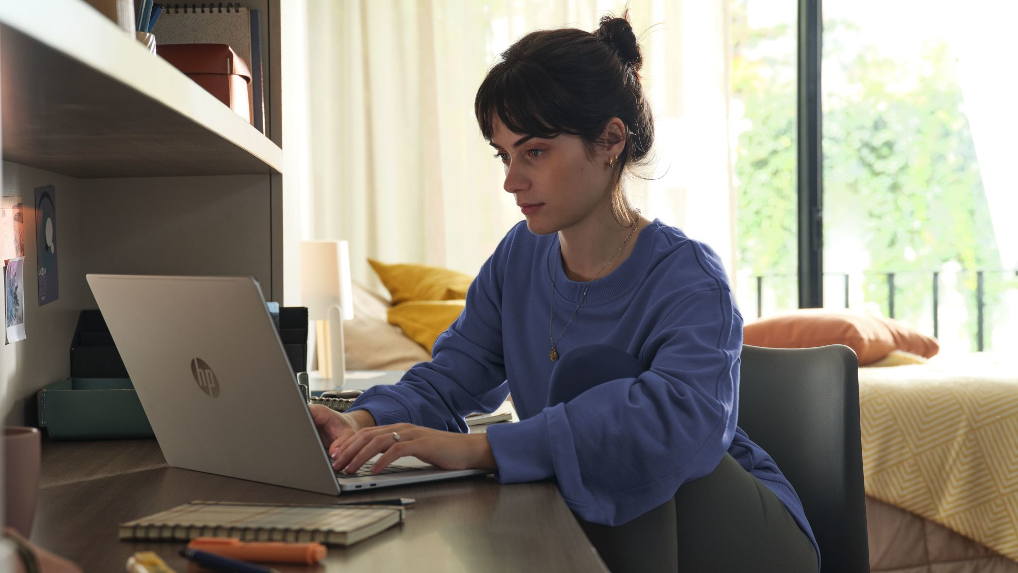 A young woman in a blue sweatshirt sits at a desk, focused on her HP laptop in a well-lit room with a cozy atmosphere.