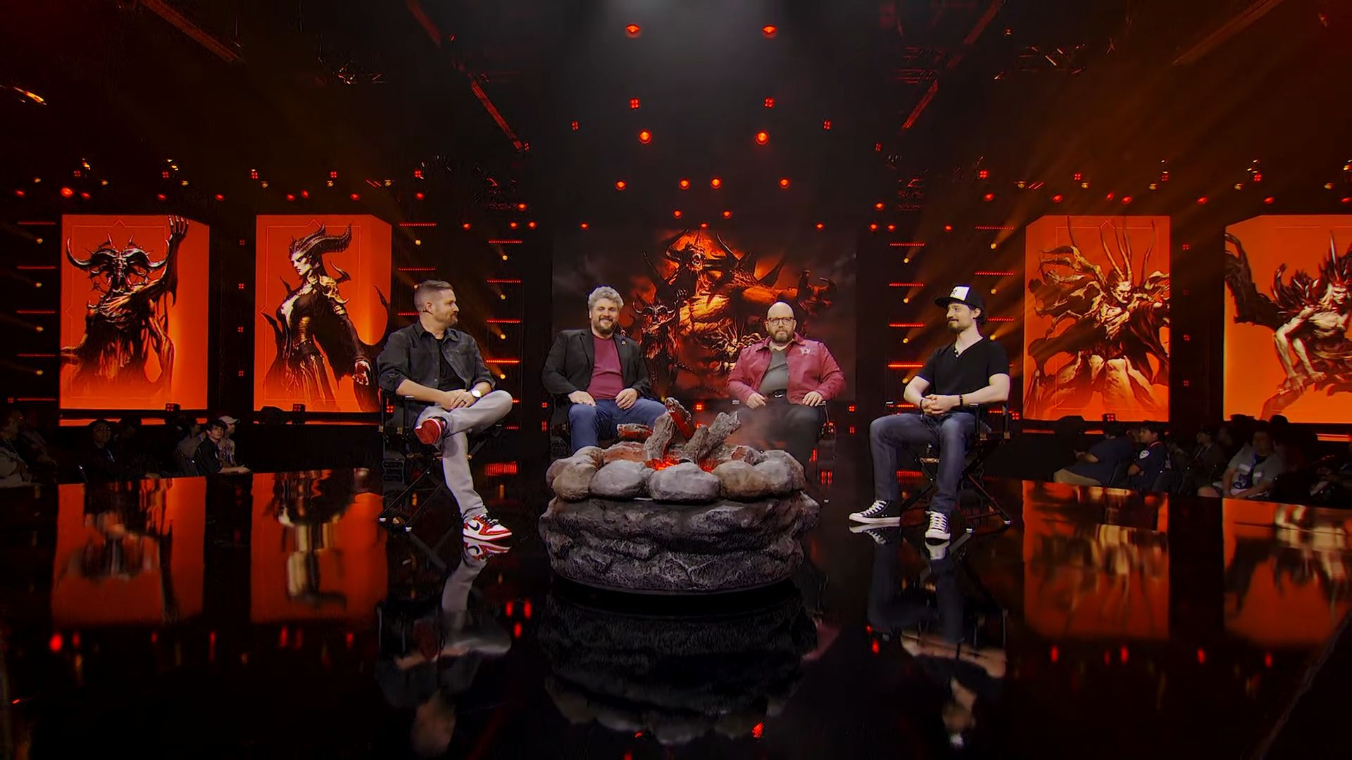 4 men sit in chairs on stage at Blizzcon, Diablo artwork displayed behind.