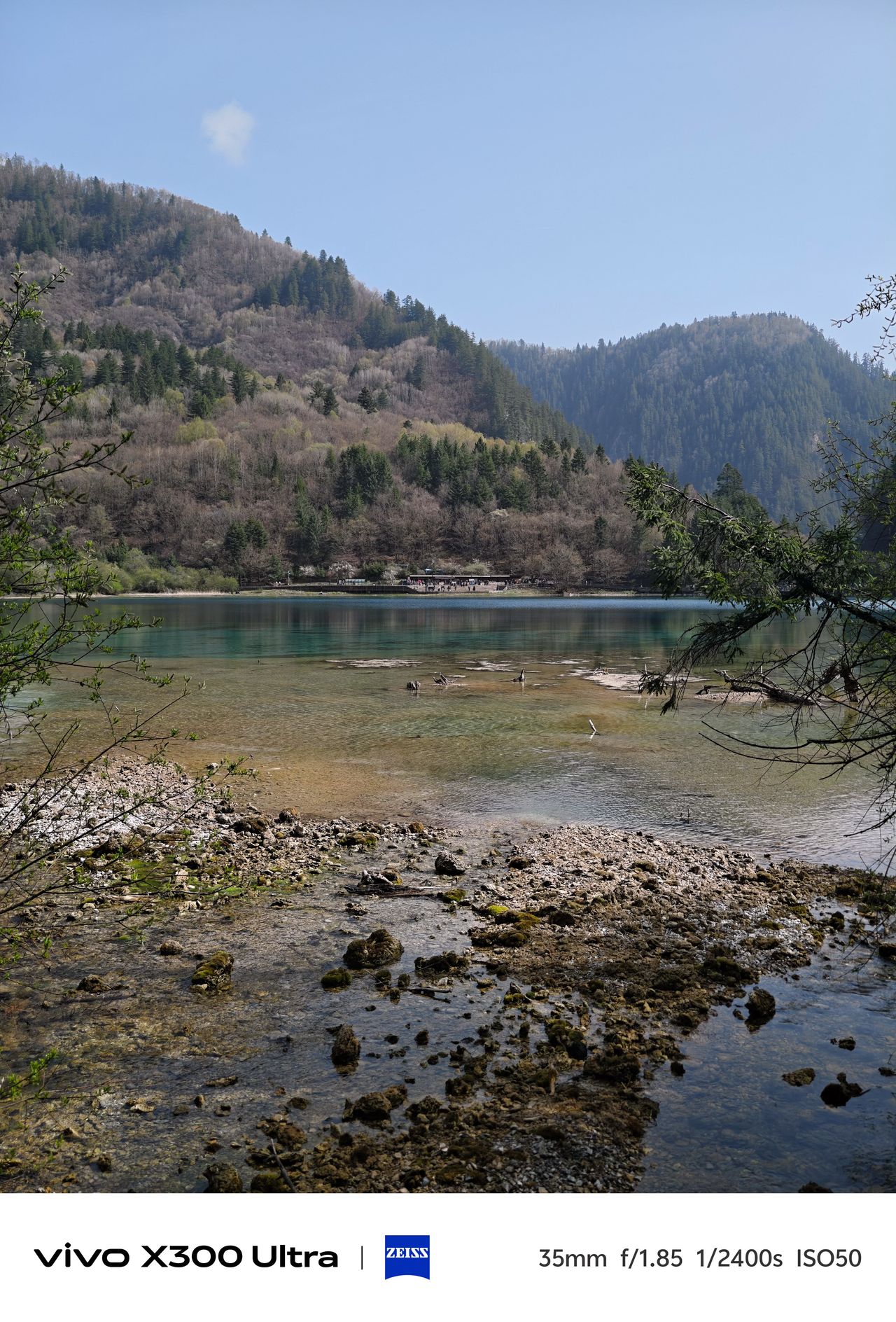 A view across a shallow, clear-water lake with a rocky shoreline and heavily forested slopes.
