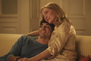 Alex (Taylor Zakhar Perez) sits on a sofa with his mother Ellen (Uma Thurman) who has her arm around his shoulders. He is leaning into her, and she is resting her head on his.