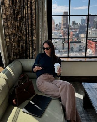 Fashion influencer Liv Perez sitting by a window with the view of New York City wearing a chic fall outfit.