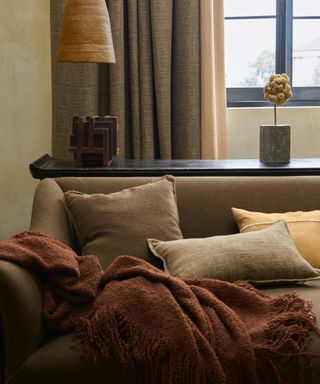 neutral living room with mocha couch with rust throw, bed cushions, mahogany console behind, taupe/grey drapes, lamps, objects