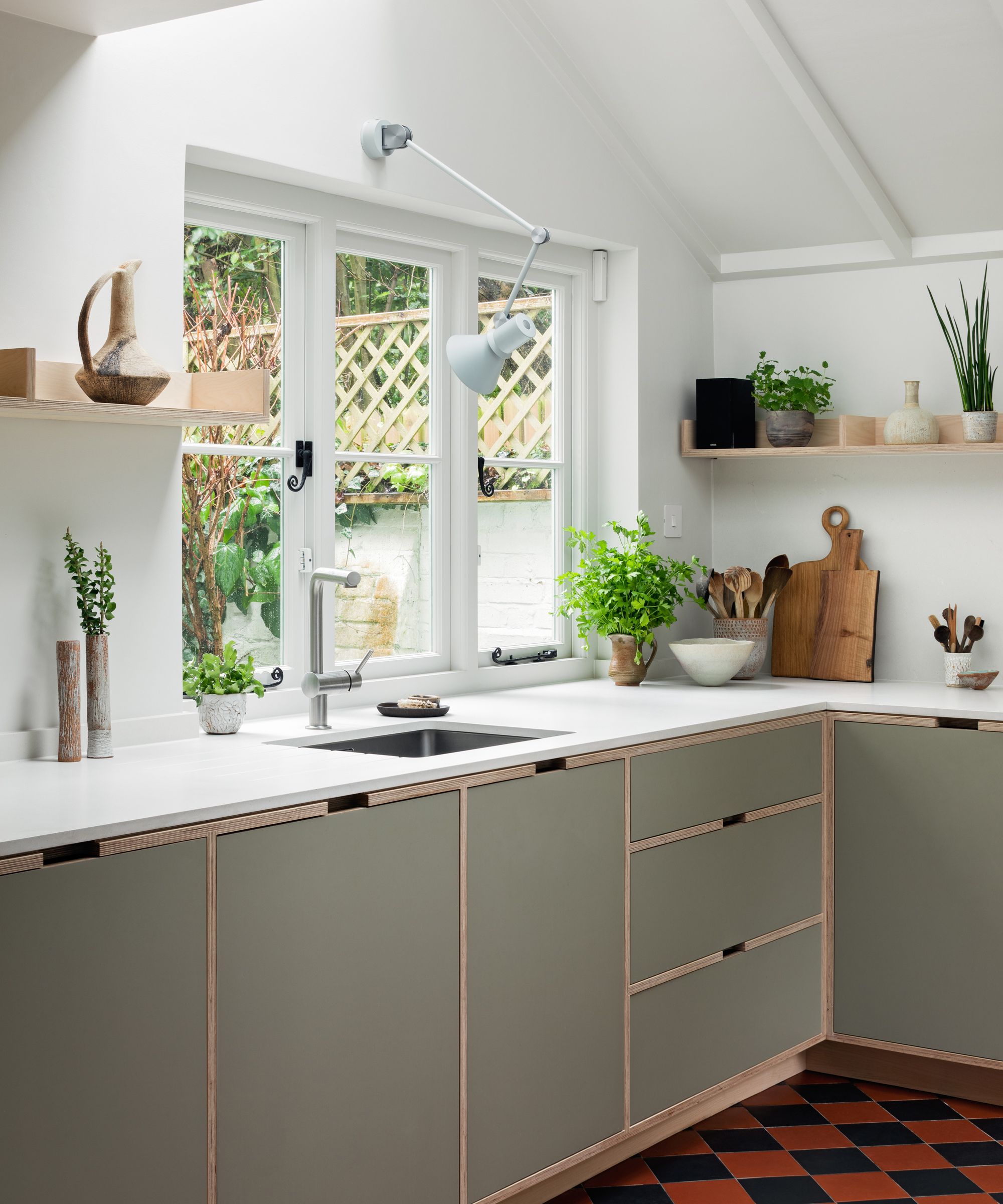 Close-up of a bright kitchen window area with sage green plywood cabinets, white countertops, and a striking black and orange checkered floor.