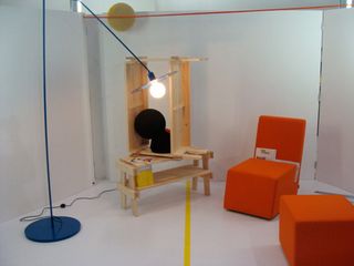 A room with white walls and floor, featuring an orange chair with matching orange side table,an oak wooden vanity table and a blue floor lamp