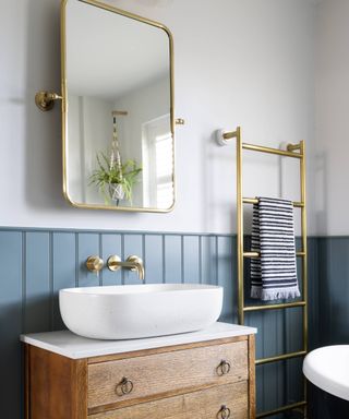 bathroom with blue wall panelling on lower half of wall, wooden chest of drawers style vanity unit, gold tower rail, mirror and taps