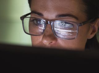 Female software developer using GitHub Copilot AI assistant on computer station with screen reflection in her spectacles.