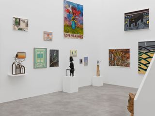 An installation view of an exhibition features multiple artworks in a white and grey gallery room, including colorful prints, paintings and sculptures.