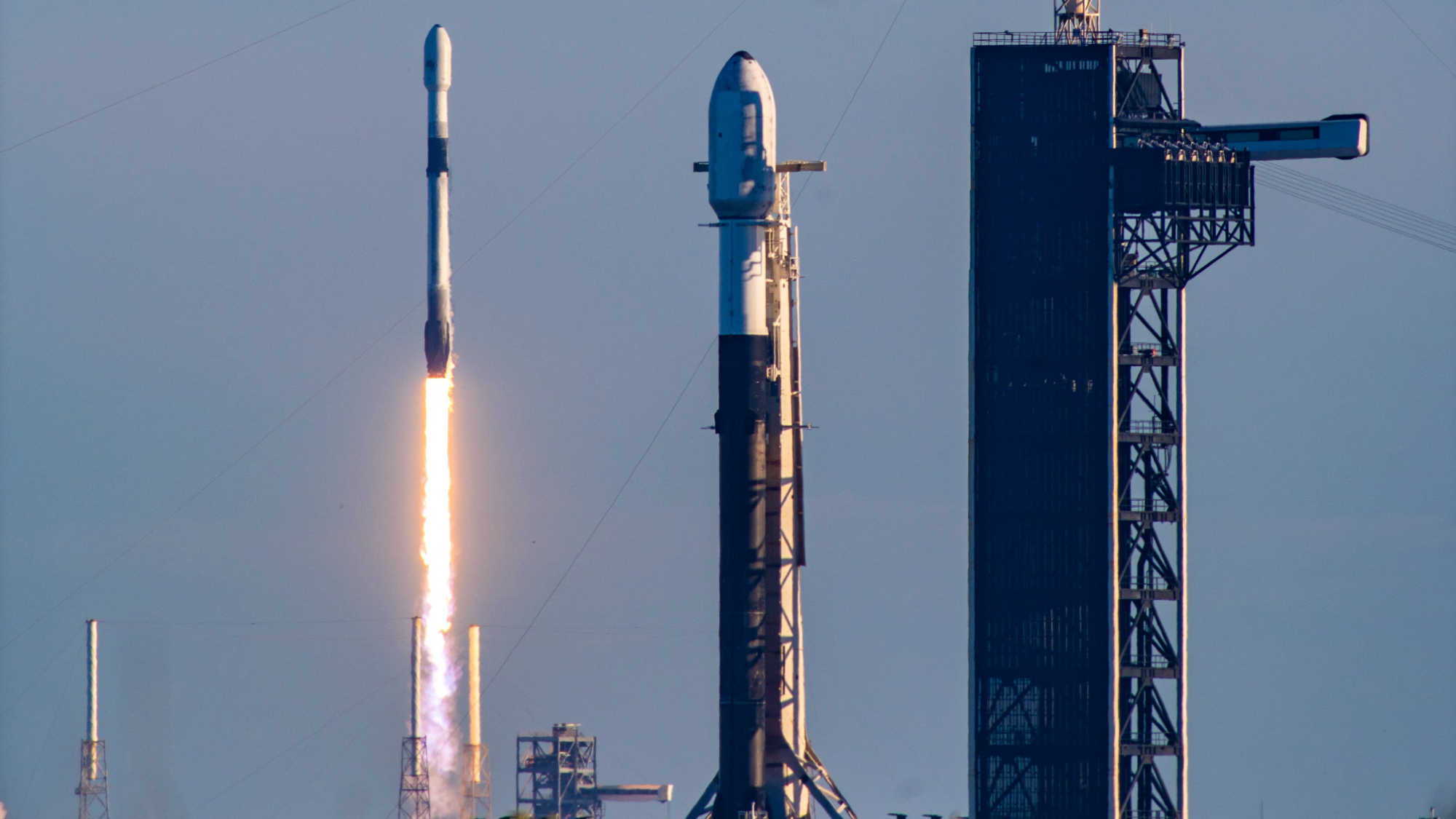 a rocket launches into a blue sky, with another rocket on a pad in the foreground