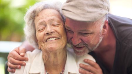 A man in his 50s hugs his elderly mother. She smiles and closes her eyes.