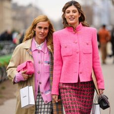 Two influencers in Paris, street styled outside of a fashion week show on a busy pavement. They are both wearing hot pink Chanel wool jackets with headbands and carrying Chanel beauty shopping bags