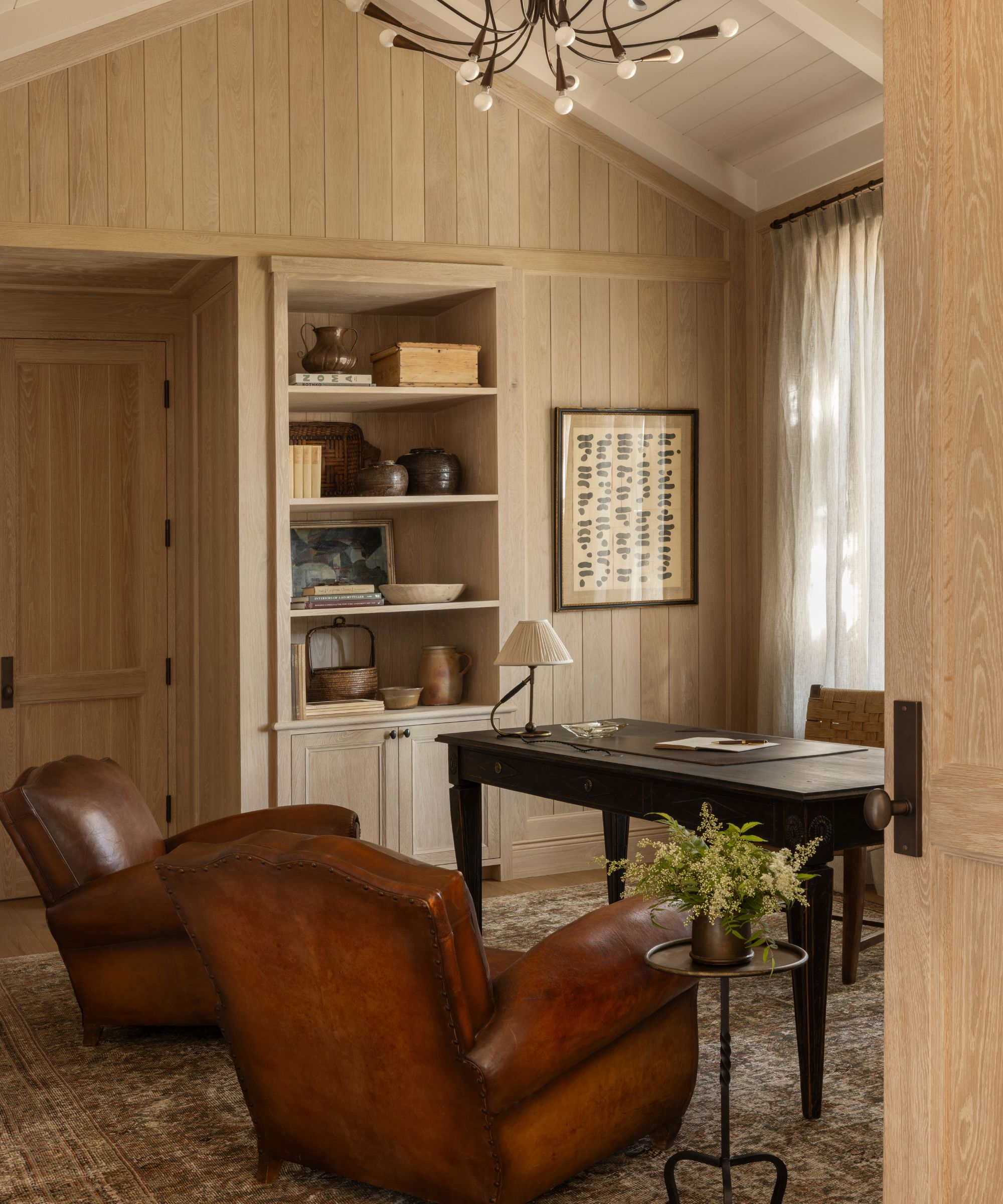 Wood-paneled home office, two leather armchairs, a wooden table, shelves of ceramics and decor, a side table with a plant on top.
