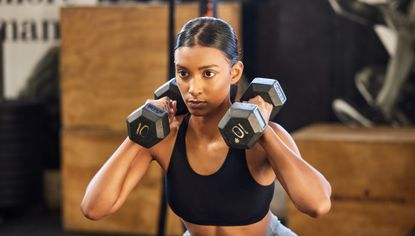 Woman in gym in squat position holding dumbbells by her shoulders. She looks focused.