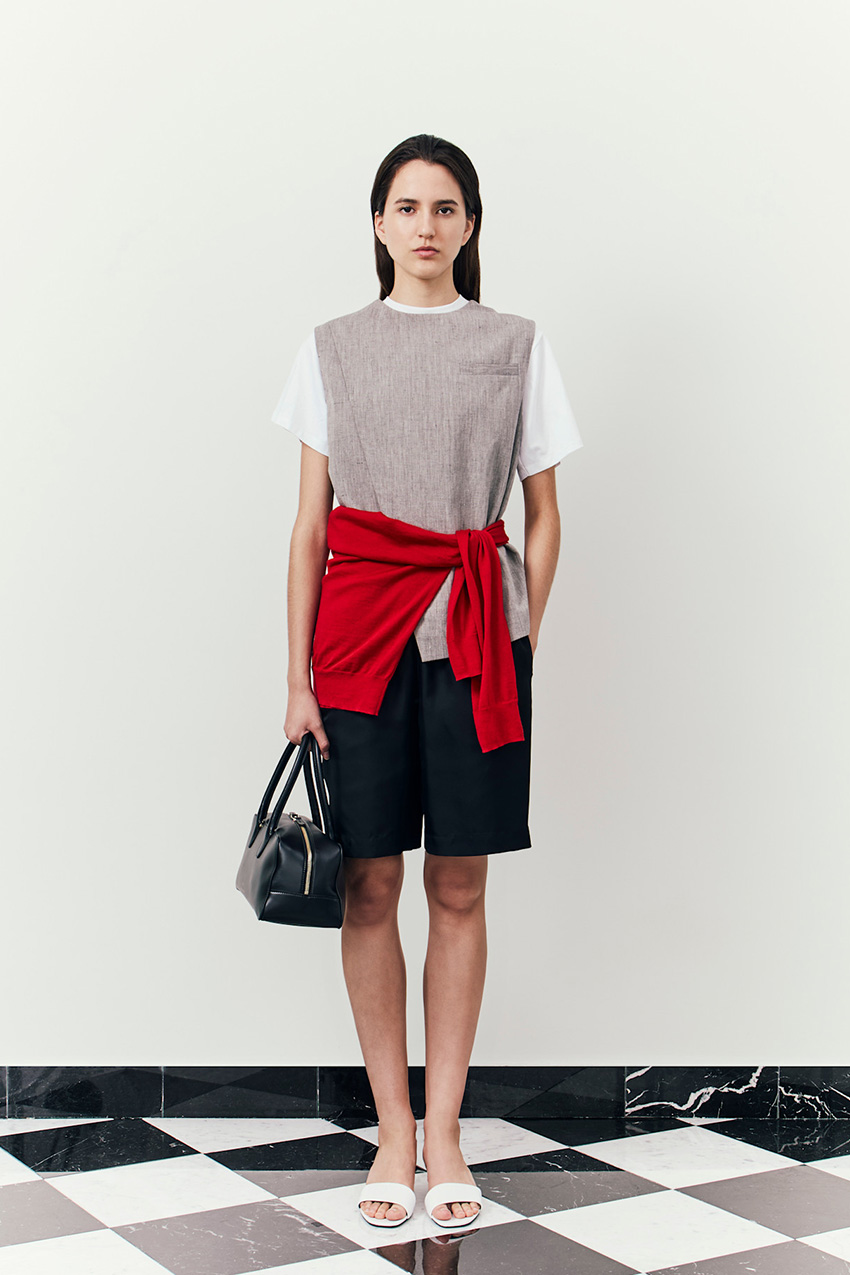Layering staples for spring 2026 is shown in a photo from Loulou De Saison's runway show, with a model walking while wearing a white t-shirt styled under a gray vest styled with a red cardigan styled around the waist, paired with black bermuda shorts, white slide sandals, and a black bowler bag