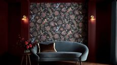 Dark red painted living room with wallpaper mural, sconces, and velvet blue curved sofa with cushion and a side table with vase holding roses