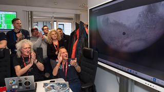 scientists applaud while looking at a flat panel TV screen displaying a image of a planet and its moon