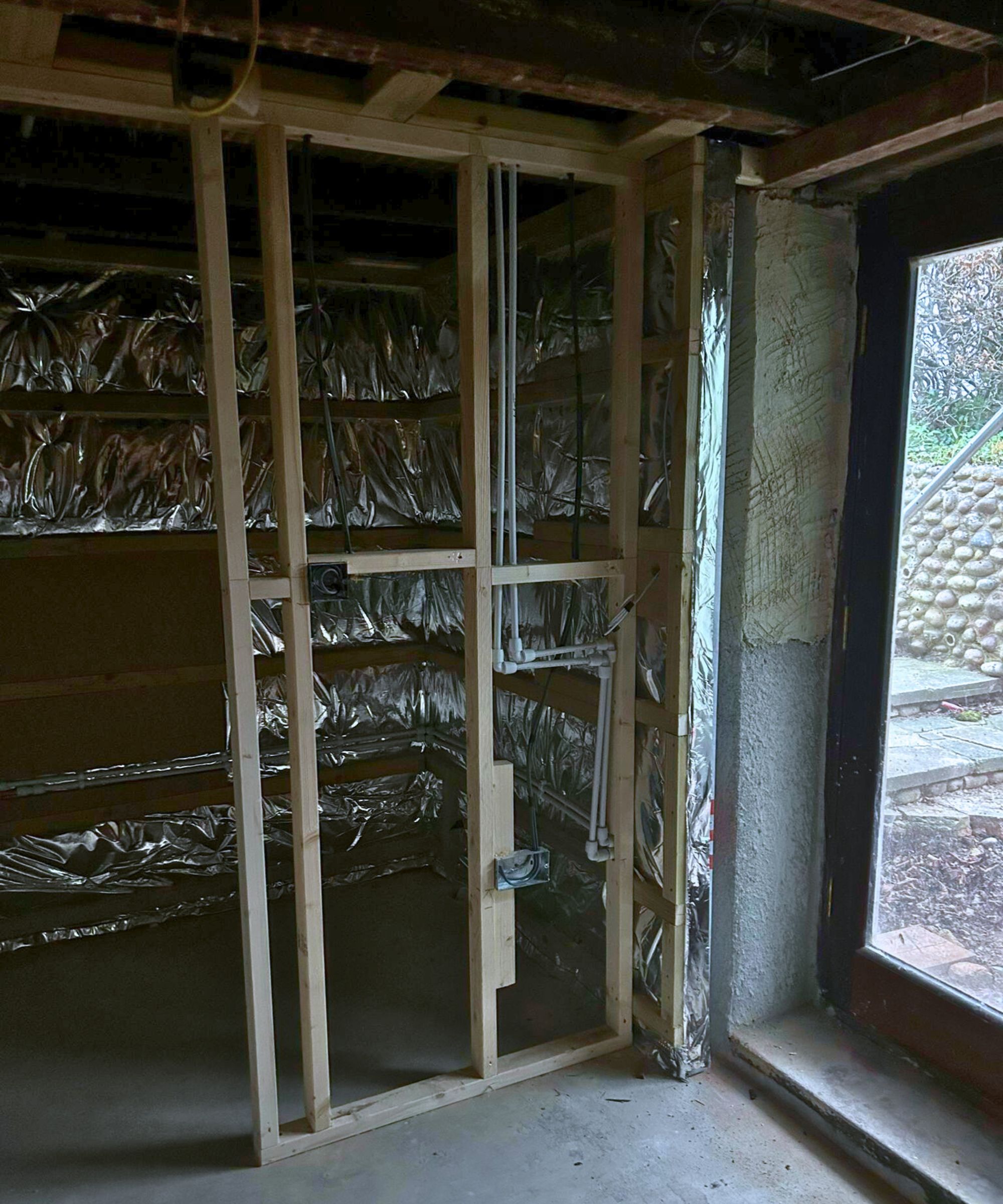 A newly framed internal wall with exposed timber studs and visible plumbing set against foil-backed insulation. The concrete floor and large glass door suggest an early-stage renovation, with raw materials and unfinished surfaces throughout the space.
