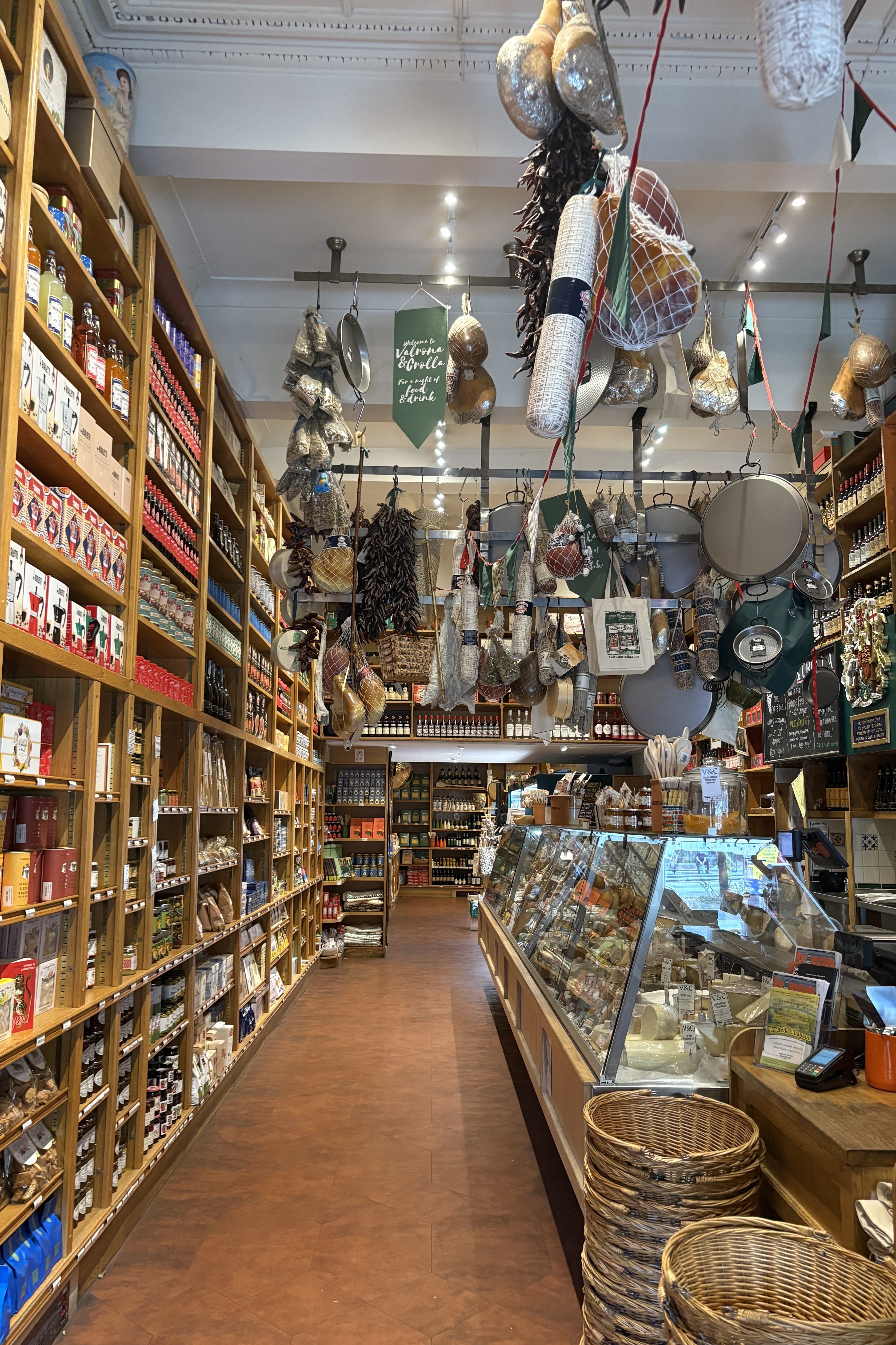 A deli stocked with all types of cheese, ham products, pickle jars, and more features a tiled terracotta floor and bright lighting.