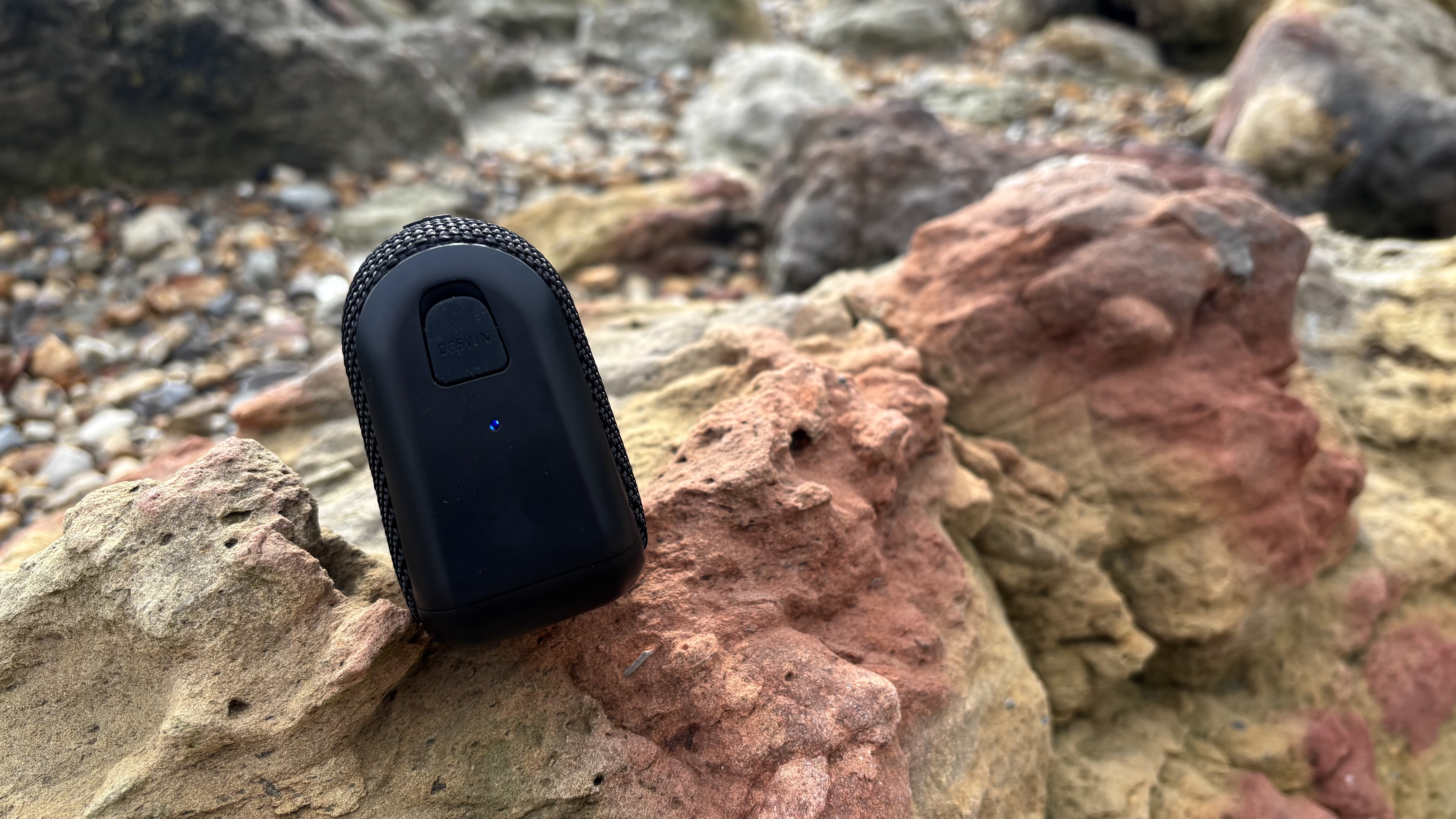 Side panel, with the blue light showing Bluetooth connection, of the black AO mini portable wireless speaker on a jagged rock.