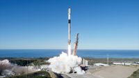 a white rocket with a black interstage lifts off from its oceanside launch pad into a clear blue sky