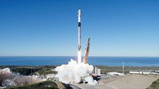 a white rocket with a black interstage lifts off from its oceanside launch pad into a clear blue sky