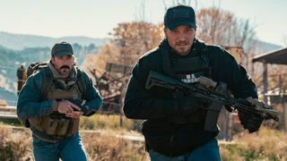 Kayce and another man run across a field holding guns