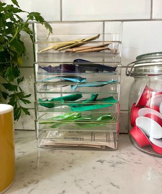 Clear plastic drawer organizers with a gold rim being used to organize tea bags on a marble kitchen counter