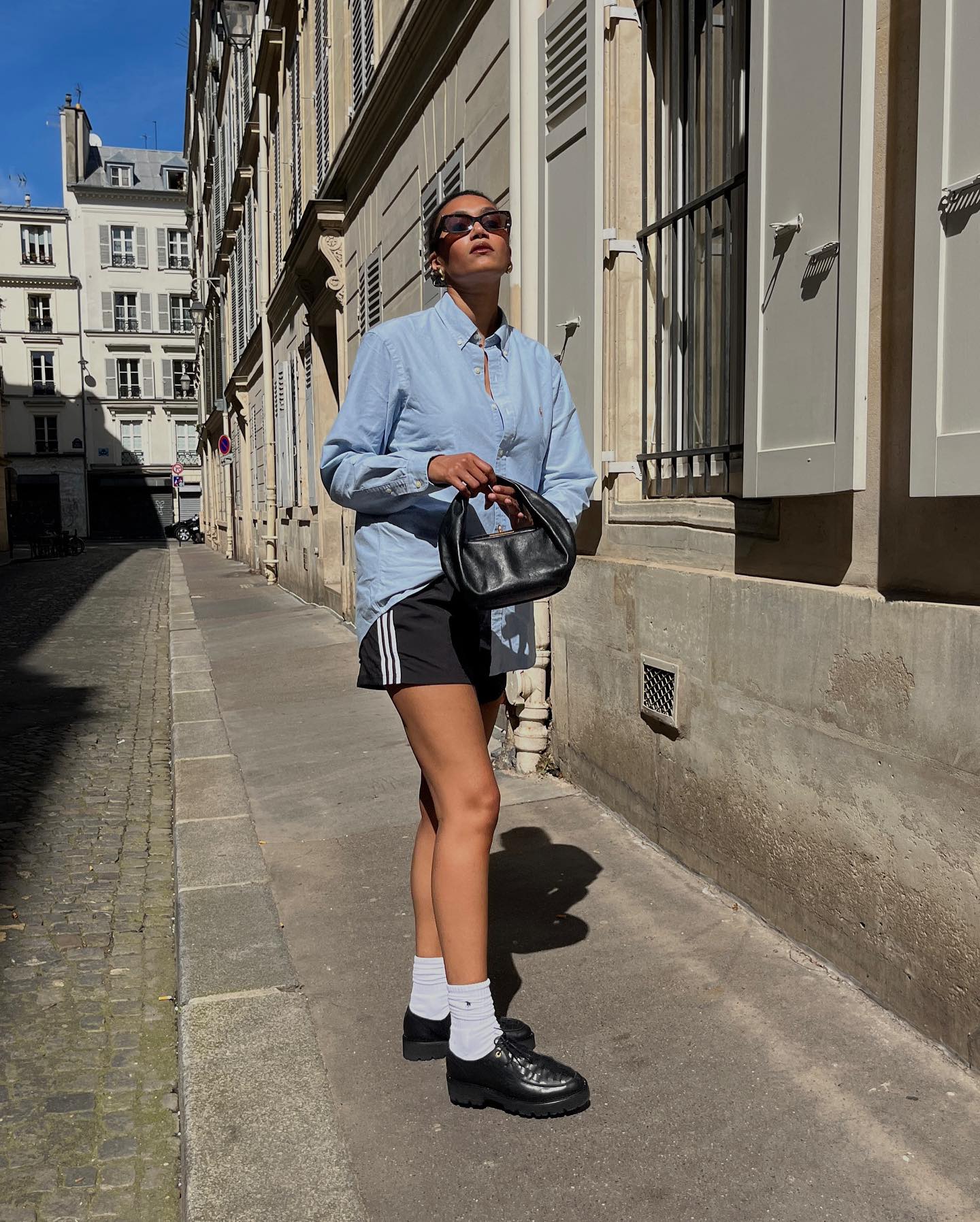 French influencer Lena Farl poses on a Paris sidewalk wearing a blue button-down shirt, black Adidas track shorts, mini black bag, white crew socks, and black oxford shoes