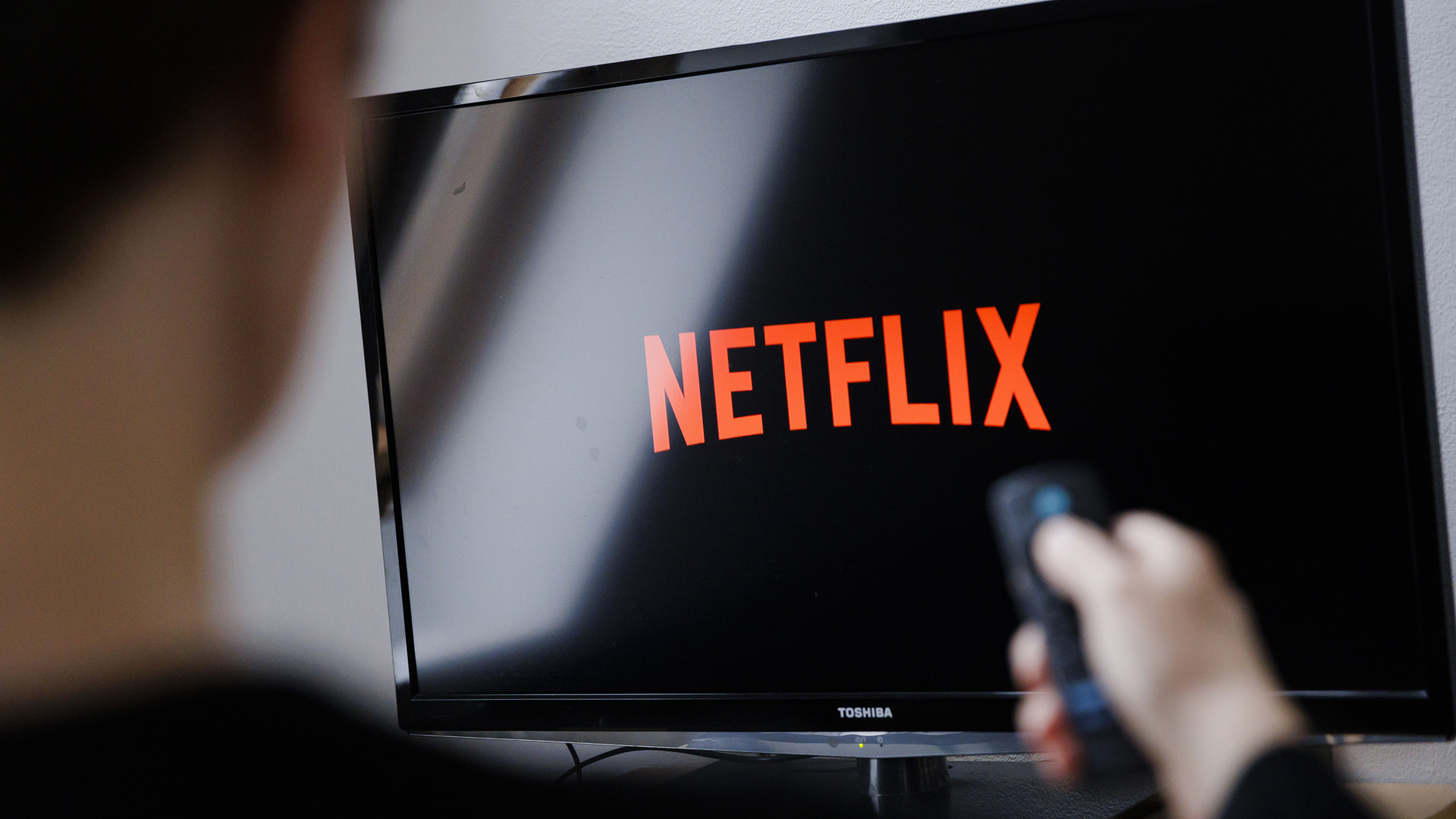 A man watches the Netflix logo on a Toshiba television with a remote control in his hand.