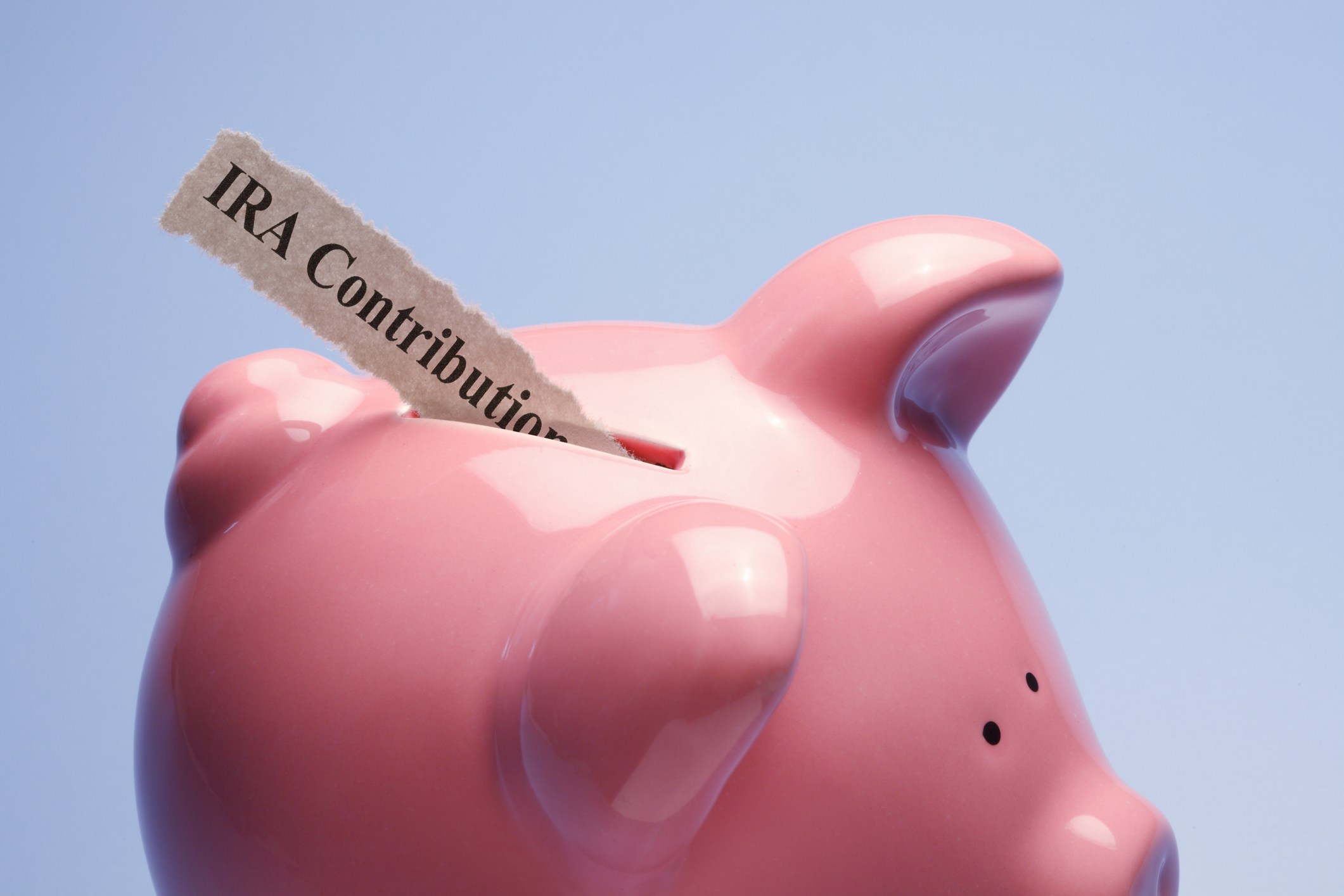&quot;IRA Contribution&quot; printed on a torn piece of paper that is inserted into the coin slot of a pink piggy bank against a blue background.