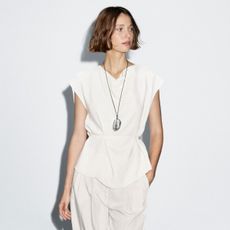 Woman wears white top, cord necklace with bead and white trousers