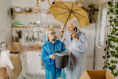 A senior couple with rain gear standing in their flooded kitchen. 