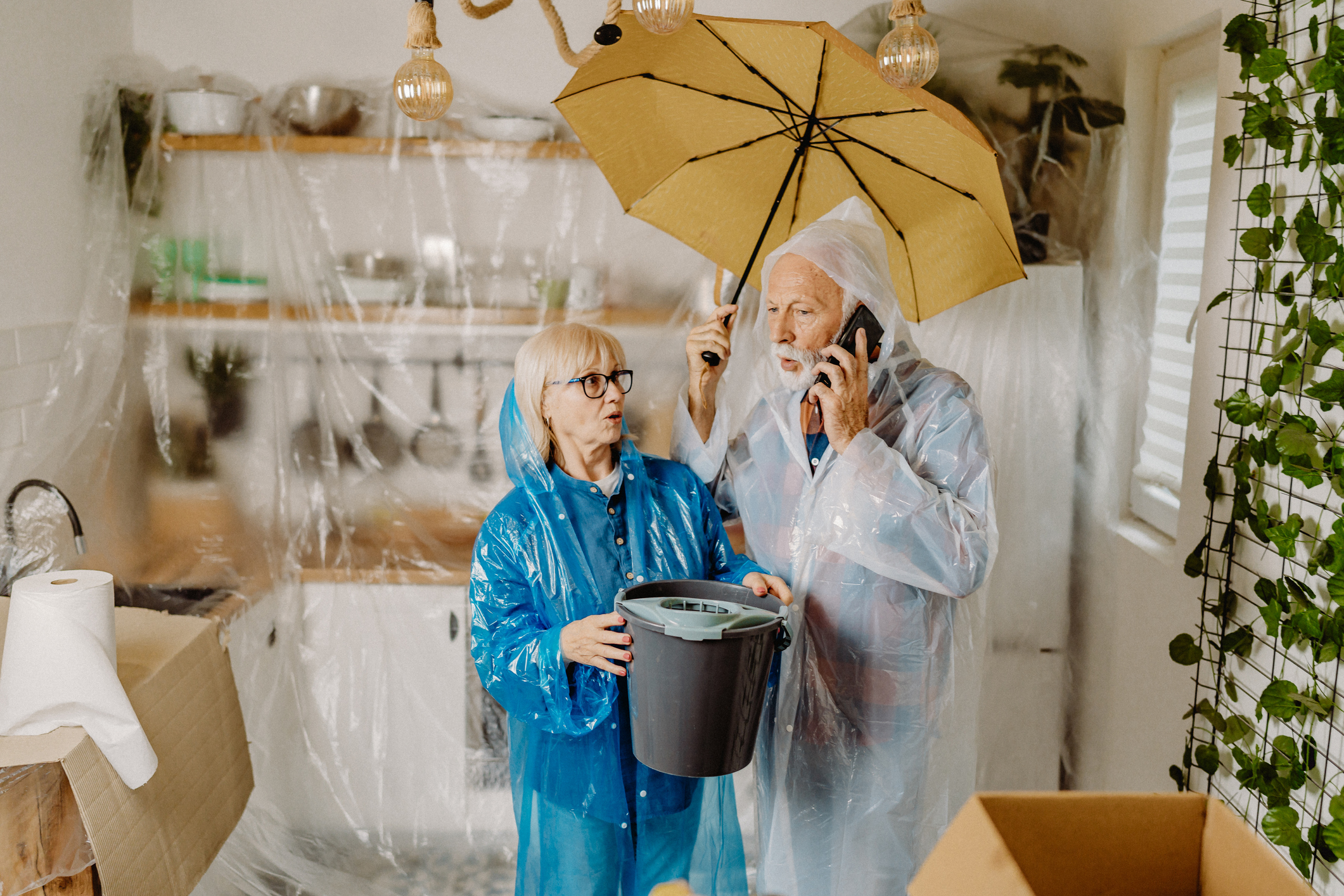A senior couple with rain gear standing in their flooded kitchen.