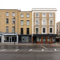 Light brown facebrick building with many windows and shops at the bottom with street parking in front of building