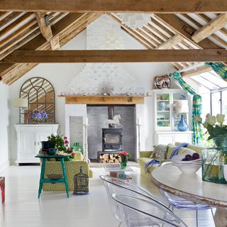 a large open farmhouse rustic style living room with white walls and fireplace, and exposed wooden beams, with green patterned sofas in the middle and a dining table with transparent chairs at the front of the image