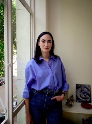 Frida Escobedo standing by a window wearing a blue shirt and jeans.
