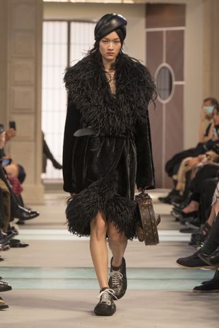 A model wearing a black fur-trim coat, black leather hat, and hiking sneakers at the Louis Vuitton F/W 25 show.