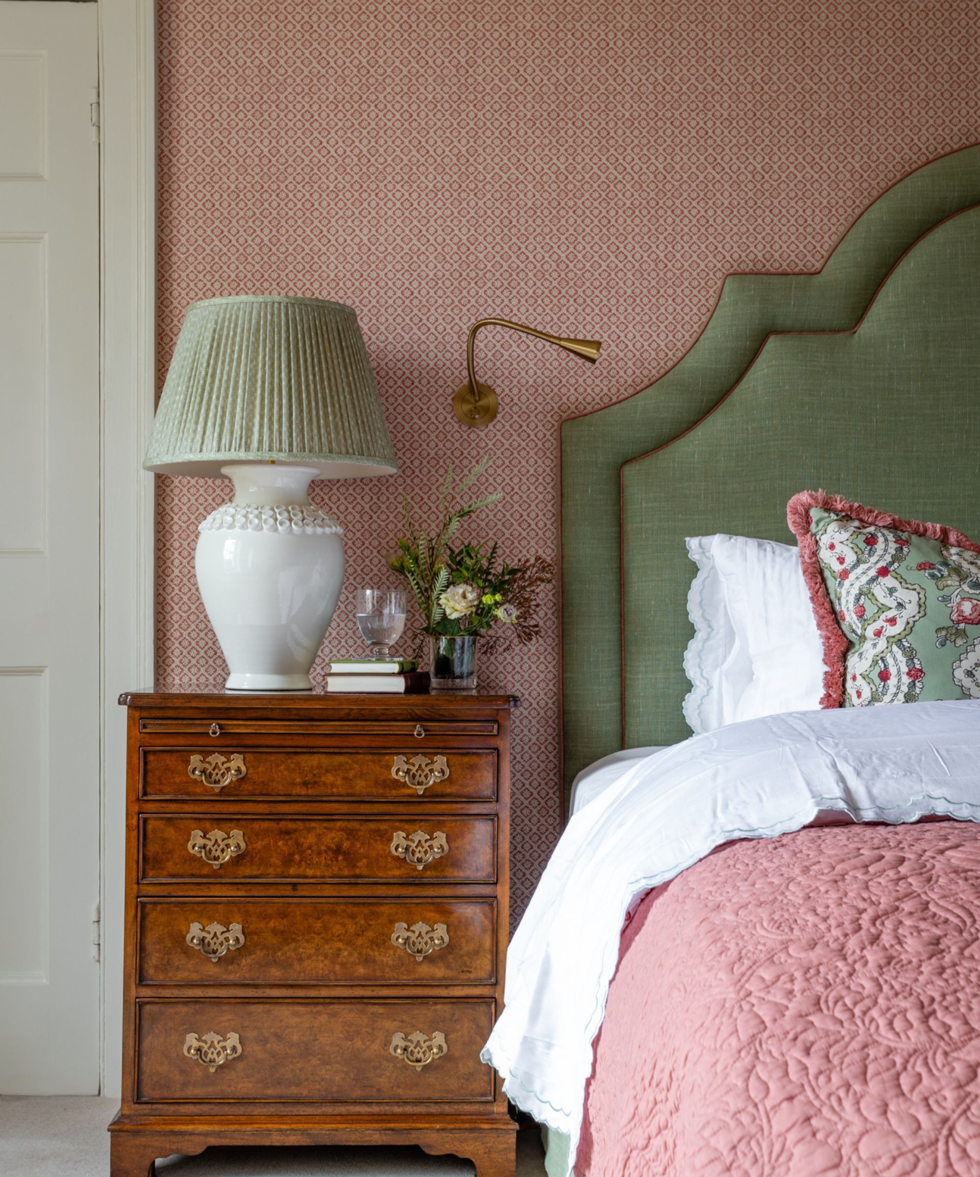 An antique bedside table with drawers, on top is a large white bedside table lamp with a green lampshade. The bed has a green headboard with pink piping and the wall has small print red wallpaper.