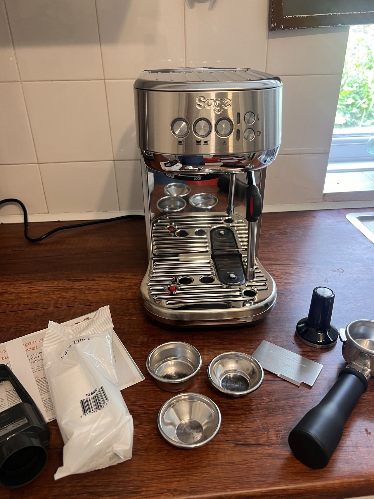 Image of a silver Sage Bambino Plus on a wooden countertop surrounded by coffee machine accessories that go with the appliance.