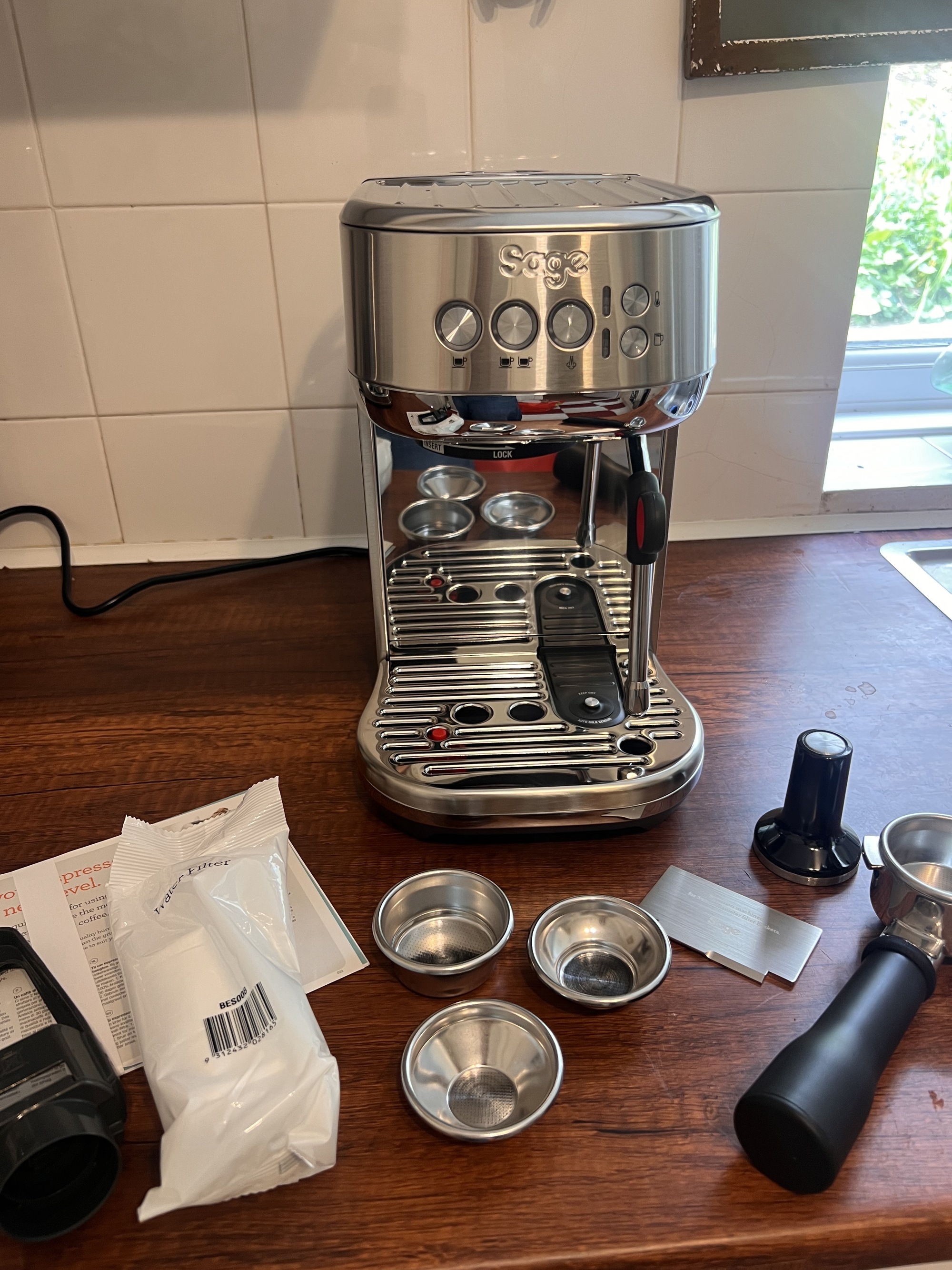 Image of a silver Sage Bambino Plus on a wooden countertop surrounded by coffee machine accessories that go with the appliance.