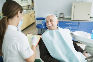 Elderly Caucasian male visiting a dentist and smiling while a young Caucasian nurse is taking notes on the patient and wearing a protective face mask, while he sits in the dentist chair