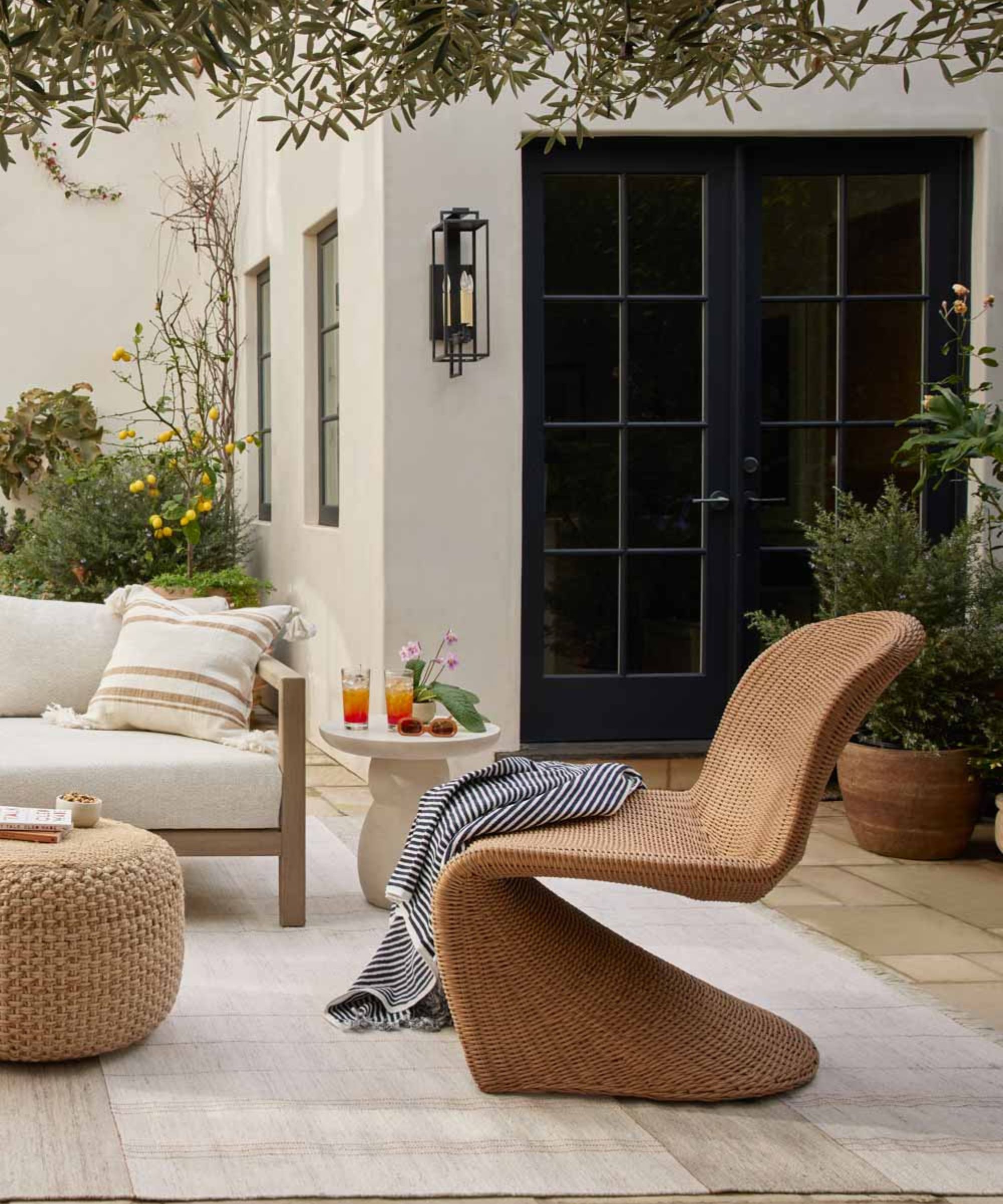 A stylish outdoor terrace featuring a sculptural, S-shaped woven lounge chair and a matching round ottoman. The space includes a neutral rug, a cream sofa with striped pillows, and a black French door in the background