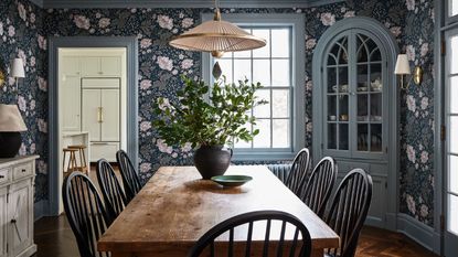 Dining room with floral blue wallpaper, blue painted windows and door frame, wood dining table and chairs, pendnat light and large vase with flowers