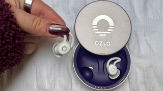 A close-up shot shows a hand holding a white Ozlo Sleepbud next to its open, circular, silver-grey charging case. The case lid features the Ozlo logo and the word "OZLO." The Sleepbuds have a small, earhook-like design. The background is a white, slightly textured fabric.