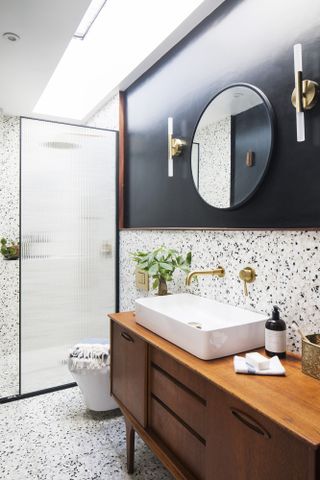 Bathroom with terrazzo wall and floor tiles, a segment of wall painted black, ribbed glass shower screen, teak sideboard turned vanity unit, brass hardware and round mirror