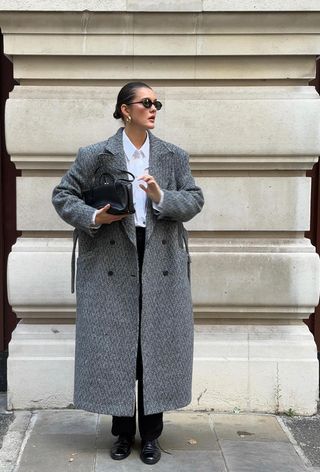 A cute work winter outfit is shown in a photo of a woman standing on the sidewalk in London wearing a long herringbone coat over a white button-down shirt with dark wash black jeans styled with a black bag, gold earrings, black sunglasses, and black oxford shoes