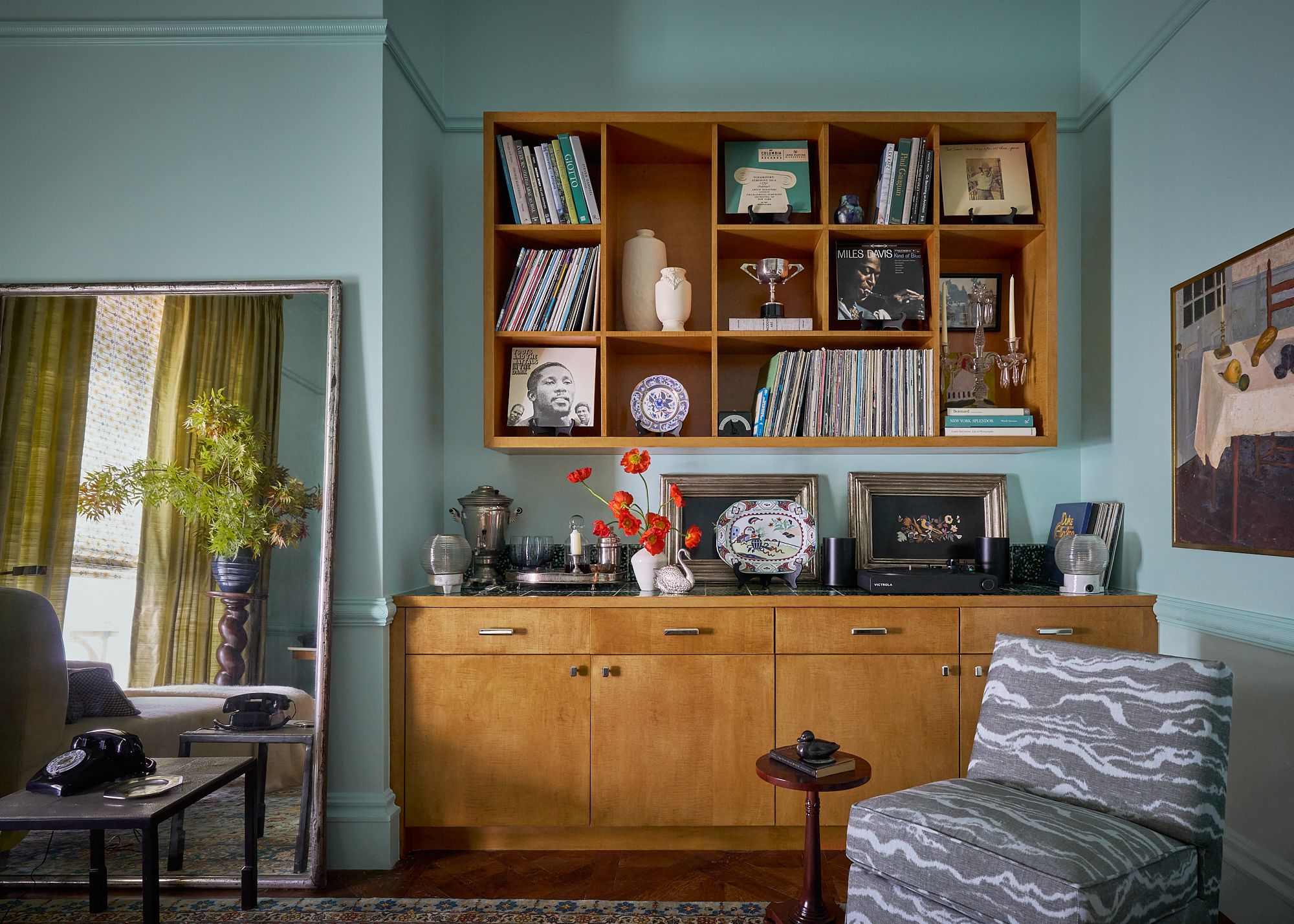 A living room with blue color-drenched walls, a large wooden cabinet with vintage decor, and a matching wooden wall unit above, with styled decor and books.