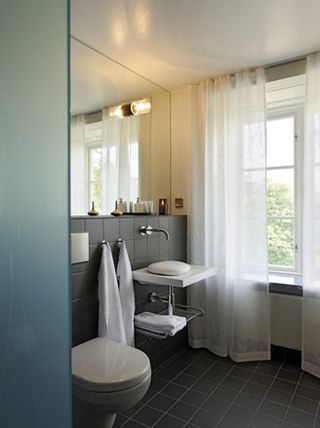 Hotel Skeppsholmen, Stockholm. A hotel bathroom with black square tiles, a rounded toilet, a white vanity, a window with white curtains and a large wall mirror.
